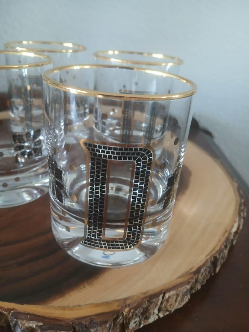 May include: A set of six vintage clear glass tumblers with gold rims and a black letter "D" design. The glasses have a mid-century modern aesthetic and are displayed on a wooden serving tray. Each glass has a capacity of approximately 240 ml.