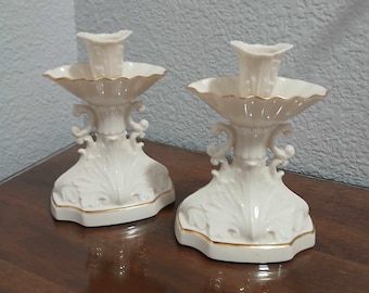 May include: A pair of ivory porcelain candle holders with gold trim. The holders have a scalloped design and are decorated with floral motifs.