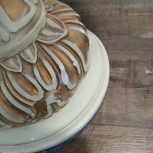 May include: Close-up of a decorative table base. The base is painted in cream and gold tones, with a detailed, ornate design. The base sits on a cream-colored, circular platform. The table base is likely part of a larger furniture piece.