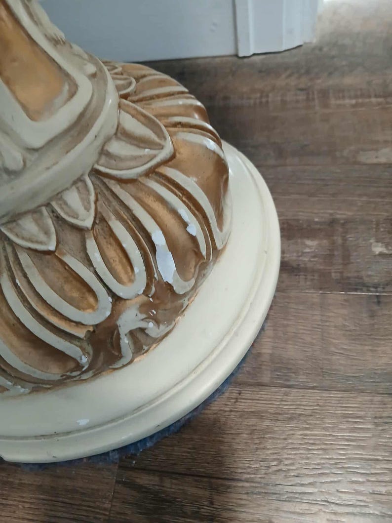 May include: Close-up of a decorative table base. The base is painted in cream and gold tones, with a detailed, ornate design. The base sits on a cream-colored, circular platform. The table base is likely part of a larger furniture piece.