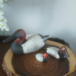 May include: Three decorative duck figurines with white, gray, black, and red coloring are displayed on a round, wooden platter. The ducks vary in size, with the largest duck measuring approximately 10 inches in length.