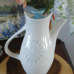 May include: White ceramic teapot with a light blue lid and handle. The teapot features a stylized sunburst design in light blue and gray. The teapot is a vintage style and is likely used for serving tea.