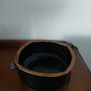 May include: A black ceramic bowl with a textured, brown rim and small handles. The interior of the bowl is dark blue. The bowl is sitting on a dark brown wooden surface.