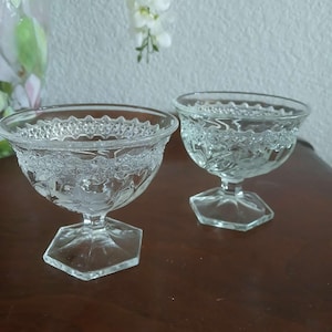 May include: Two clear glass dessert bowls with a decorative floral pattern around the rim and on the bowl. The bowls have a hexagonal base and a stem. The bowls are on a dark brown surface.