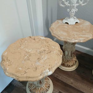 May include: Two decorative side tables with beige marble-look tops and ornate, cream-colored pedestals. One table has a candelabra on top. The tables are on a dark wood floor.