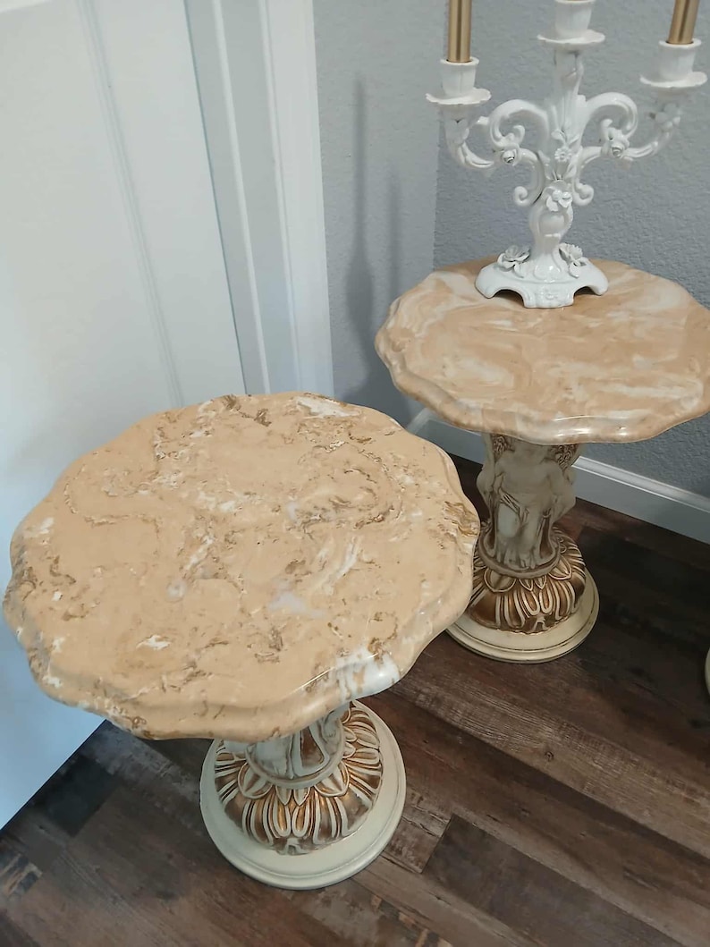 May include: Two decorative side tables with beige marble-look tops and ornate, cream-colored pedestals. One table has a candelabra on top. The tables are on a dark wood floor.