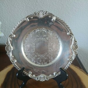 May include: A silver-colored, ornate plate with a detailed floral pattern in the center and along the rim. The plate is supported by a black stand, showcasing its decorative design. The plate is likely vintage.