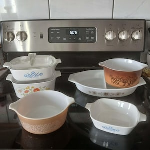 May include: A collection of vintage Pyrex baking dishes in various shapes and sizes. The dishes feature floral and geometric patterns in shades of orange, blue, and white. The image is taken in front of a stainless steel stove.