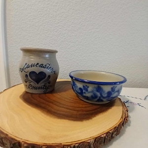 May include: A small, tan ceramic jar with the words "Lancaster County" in blue script, featuring a blue heart design. Beside it is a small blue and white patterned bowl. Both sit on a wooden slab.