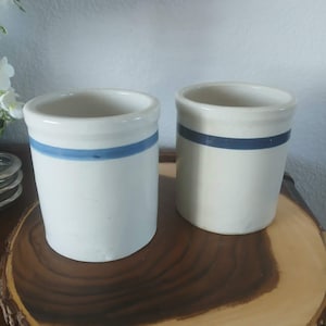 May include: Two cylindrical off-white ceramic crocks, each featuring a single horizontal blue stripe encircling the upper portion of the exterior. These containers have a smooth, matte finish and a simple, open-top design with a thick rim.