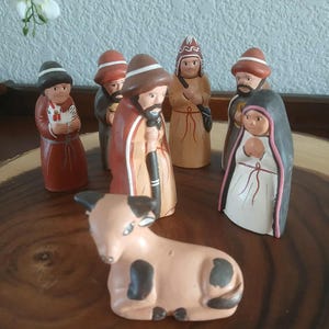 May include: A ceramic nativity scene featuring figures in various robes and hats, including a woman in a black veil and a calf with black spots. The figures are arranged on a wooden surface.