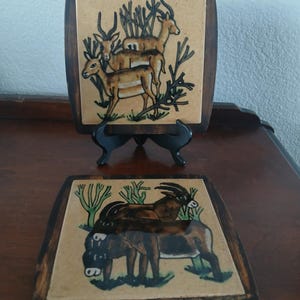 May include: Two square wooden coasters with hand-painted wildlife scenes. One coaster features three deer, the other depicts a goat and a donkey. The coasters have a dark brown wooden frame and are displayed on a dark wooden surface.