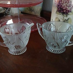 May include: A set of two clear glass sugar bowls with a ribbed design and floral pattern. The bowls have a footed base and a handle on one side.