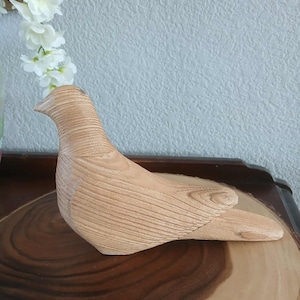 May include: A wooden bird sculpture with a natural wood grain finish. The bird is perched on a wooden surface, with white flowers in the background. The sculpture is a decorative item.
