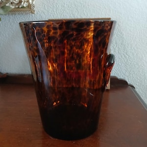 Puede incluir: Un jarrón alto de vidrio marrón con un patrón de carey. El jarrón tiene una abertura ancha y una forma ligeramente curvada. Cuenta con un pequeño asa en el lateral. El jarrón se utiliza probablemente con fines decorativos, como para sostener flores u otros objetos.