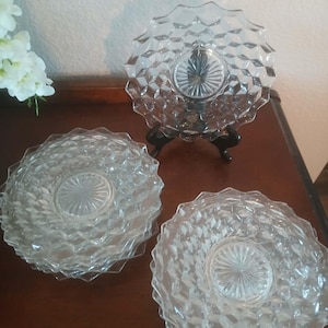 May include: Three clear glass plates with a diamond pattern and a starburst center. One plate is displayed on a black stand. The plates are on a dark brown surface.