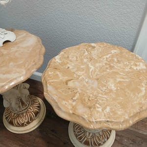 May include: Two beige marble-topped side tables. The tables have a scalloped edge and sit on a cream-colored pedestal base with gold accents. The bases have a detailed, carved design. The tables are on a wood floor.