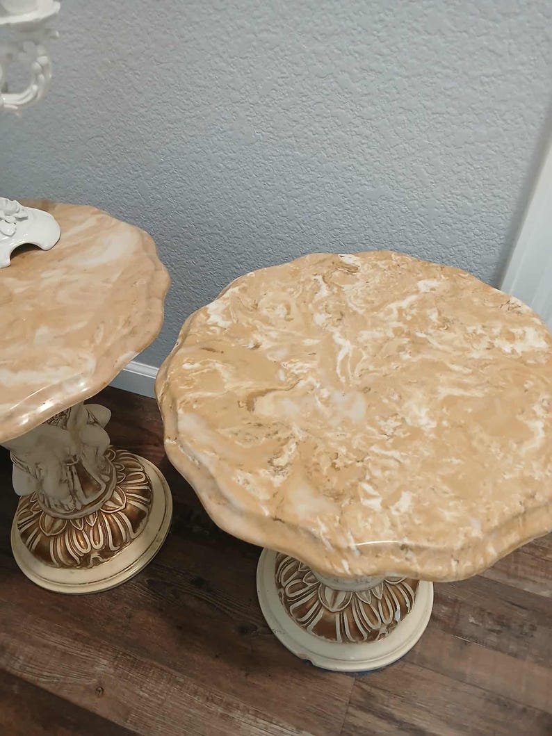 May include: Two beige marble-topped side tables. The tables have a scalloped edge and sit on a cream-colored pedestal base with gold accents. The bases have a detailed, carved design. The tables are on a wood floor.
