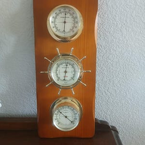May include: A wooden weather station with three brass-rimmed gauges. The top gauge is a thermometer, the middle is a barometer with a ship wheel design, and the bottom is a humidity gauge. The wood has a warm, reddish-brown tone.