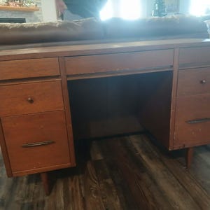 May include: A vintage wooden desk with a warm brown finish. It features multiple drawers with round and bar-shaped handles, and a central open space for a chair. The desk has tapered legs and a classic mid-century modern design.