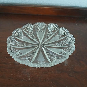 May include: Clear, cut-glass dish with a starburst design. The dish has a scalloped edge and intricate geometric patterns radiating from the center. The glass is transparent, allowing light to pass through, and sits on a dark wooden surface.