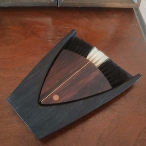May include: A small, wooden dustpan and brush set. The dustpan is black and angled, with a curved edge. The brush is triangular, with a dark wood handle and white bristles. A copper-colored accent dot is on the handle.