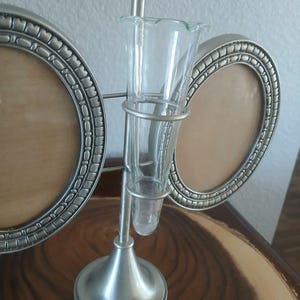 May include: A silver-toned metal photo frame with two oval openings and a clear glass bud vase in the center. The vase is held by a metal frame and has a flared top. The frame sits on a wooden surface.