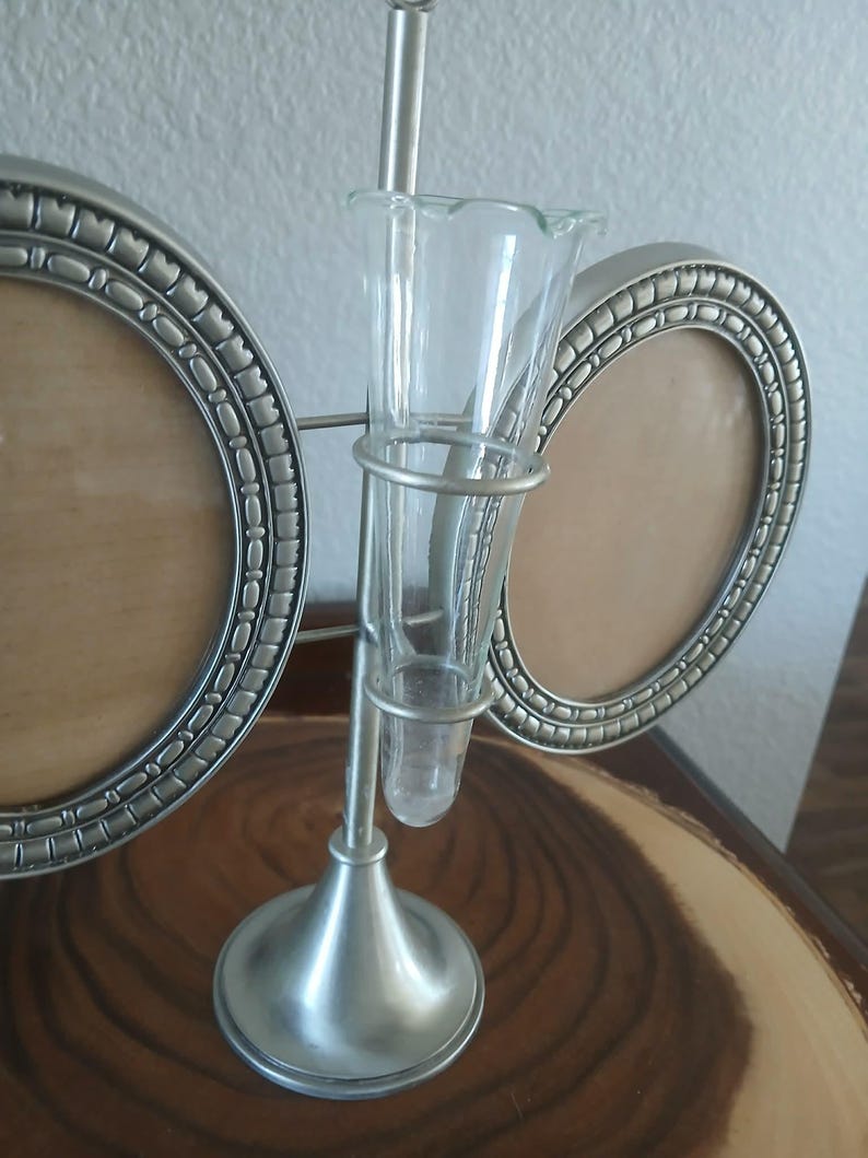 May include: A silver-toned metal photo frame with two oval openings and a clear glass bud vase in the center. The vase is held by a metal frame and has a flared top. The frame sits on a wooden surface.