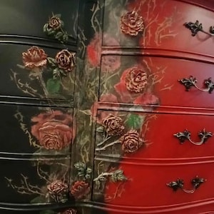 May include: A two-toned dresser, black on the left and red on the right, adorned with rose-shaped embellishments and floral artwork. The drawers feature ornate, dark-colored handles. The overall design is a blend of gothic and romantic styles.