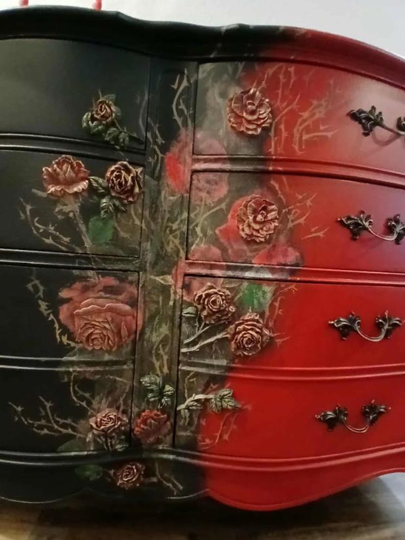May include: A two-toned dresser, black on the left and red on the right, adorned with rose-shaped embellishments and floral artwork. The drawers feature ornate, dark-colored handles. The overall design is a blend of gothic and romantic styles.