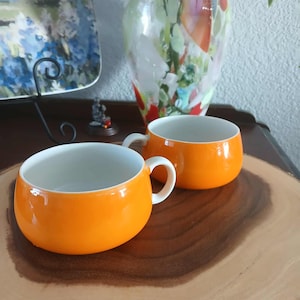 May include: Two bright orange ceramic mugs with white interiors and curved handles. The mugs are on a wooden surface, with a decorative vase and a plate in the background. The mugs have a rounded shape.