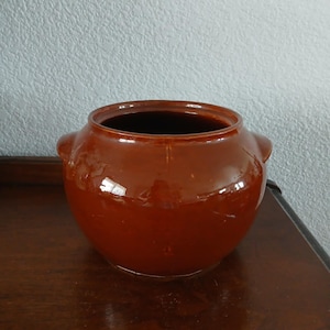 May include: A glazed, brown ceramic pot with two small handles on the sides. The pot has a rounded body and a wide opening. The interior is dark, and the exterior has a glossy finish. The pot is sitting on a wooden surface.