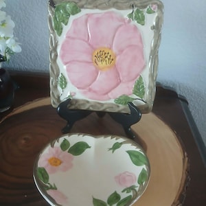 May include: A square ceramic decorative plate with a pink flower design and a heart-shaped dish with a similar floral pattern. The plate is supported by a black stand, and the dish is on a wooden surface. The floral design includes pink petals, yellow centers, and green leaves.