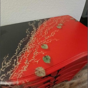 May include: A red and black painted dresser with a decorative design. The top of the dresser is split diagonally, with the left side black and the right side red. A thorny vine design and green leaves are painted on the top.