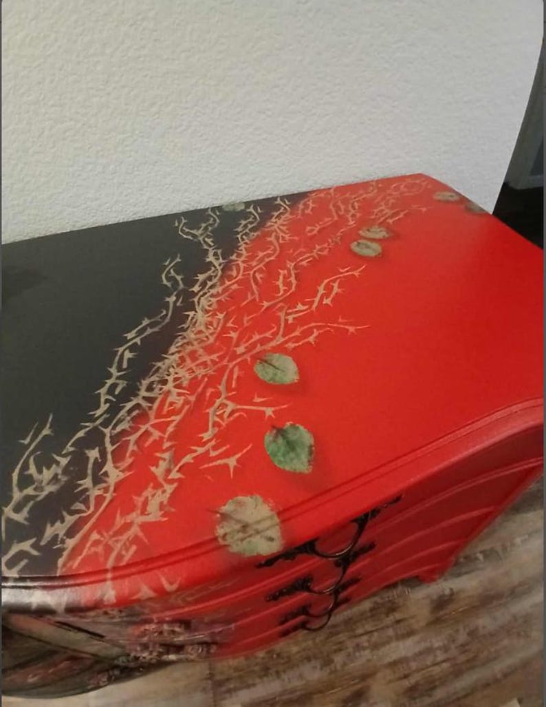 May include: A red and black painted dresser with a decorative design. The top of the dresser is split diagonally, with the left side black and the right side red. A thorny vine design and green leaves are painted on the top.