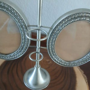 May include: A silver-colored, double photo frame with a decorative, oval design. The frame has a central support with a decorative base and two circular holders for photos. The frame is made of metal and sits on a wooden surface.