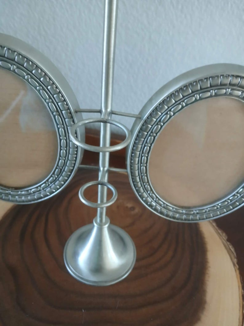 May include: A silver-colored, double photo frame with a decorative, oval design. The frame has a central support with a decorative base and two circular holders for photos. The frame is made of metal and sits on a wooden surface.