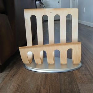 May include: A wooden magazine rack with a silver base. The rack has five slots for magazines and is made of light wood.