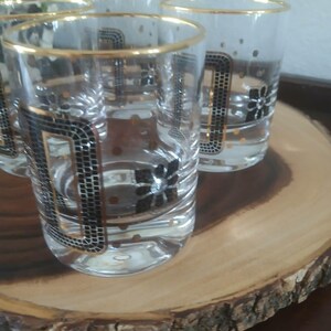 May include: A set of six clear drinking glasses with gold rims and black and gold geometric patterns. The glasses have a mosaic-like design and floral accents. The glasses are displayed on a wooden serving tray.