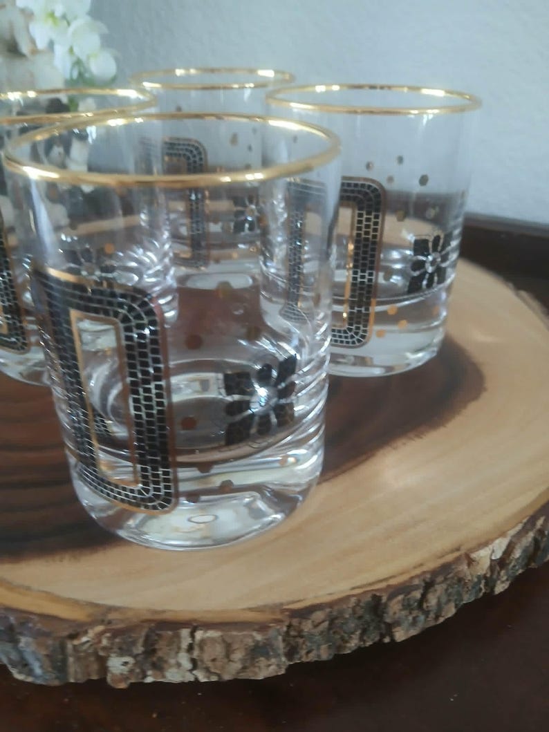 May include: A set of six clear drinking glasses with gold rims and black and gold geometric patterns. The glasses have a mosaic-like design and floral accents. The glasses are displayed on a wooden serving tray.