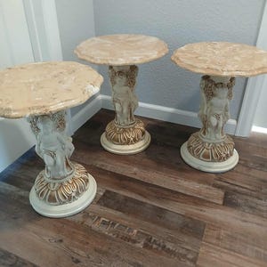 May include: Three decorative pedestal tables with round, light-colored tops. Each table features a cherub figure supporting the top, with a detailed, ornate base. The tables are cream and beige in color, and are placed on a wood-look floor.