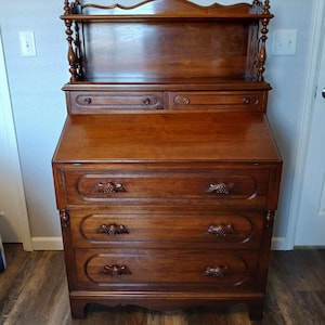 May include: A dark wood antique secretary desk with a drop-down writing surface, four drawers, and a top shelf. The desk has intricate carvings and a curved top.