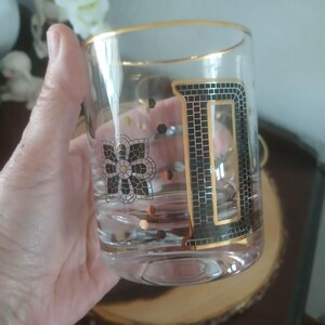 May include: A clear glass tumbler with a gold rim and a large, black and gold letter "D" design. The glass also features a floral design and gold accents. The glass is held in a hand.