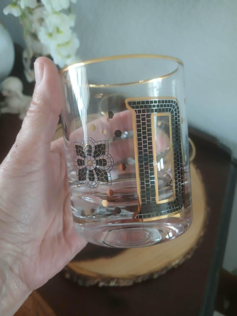 May include: A clear glass tumbler with a gold rim and a large, black and gold letter "D" design. The glass also features a floral design and gold accents. The glass is held in a hand.