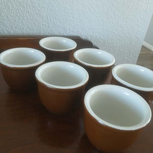 May include: Six small, brown ceramic ramekins with white interiors and a glossy finish. The ramekins are arranged on a dark wooden surface, showcasing their rounded shape and simple design. Ideal for serving individual portions of desserts or appetizers.