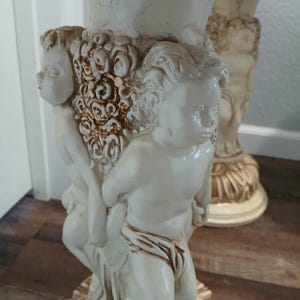 May include: Off-white decorative table leg featuring cherubic figures and floral accents. The leg has a detailed, sculpted design with a cream-colored finish and subtle gold highlights, showcasing classical artistry.