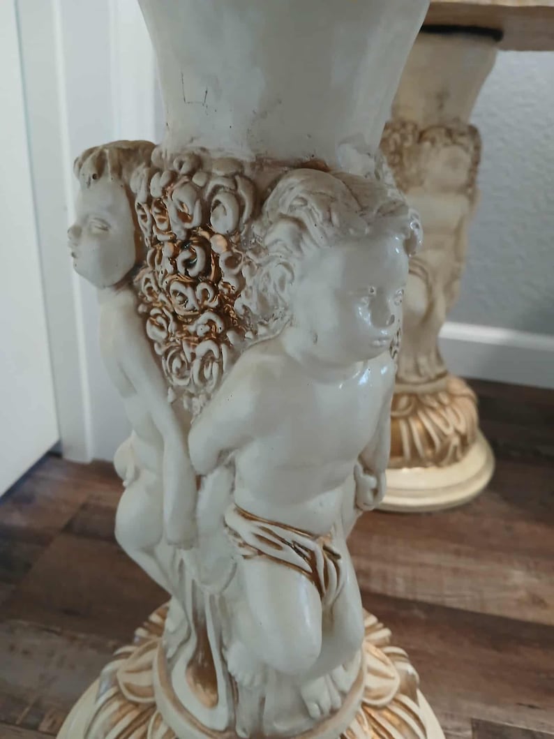 May include: Off-white decorative table leg featuring cherubic figures and floral accents. The leg has a detailed, sculpted design with a cream-colored finish and subtle gold highlights, showcasing classical artistry.