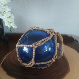 May include: A spherical, deep blue glass float encased in a decorative, knotted tan rope netting. The glass orb features a smooth, reflective surface and is secured by a thick, braided rope handle attached to one side of the netting structure.