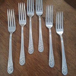 May include: Six silver forks with floral detailing on the handles are arranged on a wooden surface. The forks have four tines and a classic design, suitable for formal dining. The silverware is displayed in a neat, organized manner.