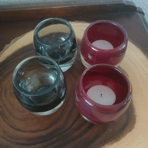 May include: Four glass votive candle holders. Two are clear, and two are a deep red color. Each red holder contains a white candle. The candle holders are arranged on a wooden surface.